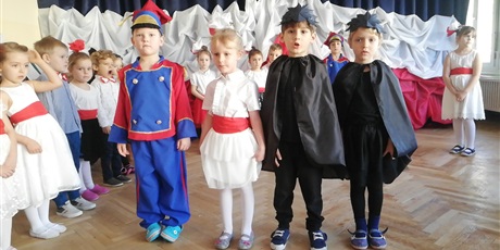 Powiększ grafikę: występ dzieci z okazji 11 listopada na tle flagi biało czerwonej.chłopiec przebrany jest w strój ułana dwie osoby przebrane za postaci ptaków czarnych wron i dziewczynka w stroju białym