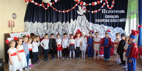 Powiększ grafikę: W tle grupa dzieci w wieku przedszkolnym ubranyc w galowe stroje biorących udział w przedsztwieniu z okazji 11 listopada