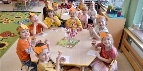 Powiększ grafikę: Na zdjęciu dzieci siedzą wokół dwóch stolików. Na głowach mają pomarańczowe papierowe opaski. Jedzą babeczki z pomarańczowym lukrem. dzieci uśmiechają się.