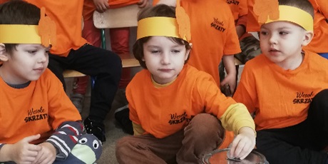 Powiększ grafikę: dziecko siedzi na podłodze ,w slali przedszkolnej w pomarańczowej koszulce z opaską ze słoniem na głowie wrzuca do metalowej puszki monetę