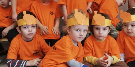 Powiększ grafikę: dziecko siedzi na podłodze ,w slali przedszkolnej w pomarańczowej koszulce z opaską ze słoniem na głowie wrzuca do metalowej puszki monetę