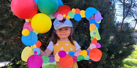 Powiększ grafikę: Na zdjęciu dziecko w wieku przedszkolnym na tle kolorowej ramki ozdobionej papierowymi kropkami i balonami.