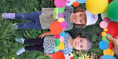 Powiększ grafikę: Na zdjęciu dzieci w wieku przedszkolnym na tle kolorowej ramki ozdobionej papierowymi kropkami i balonami.