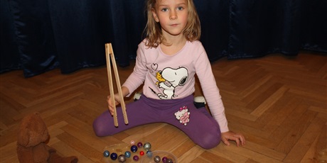 Powiększ grafikę: Na zdjęciu dziecko w wieku przedszkolnym układające szklane kolorowe kulki drewnianymi szczypcami.