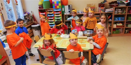 Powiększ grafikę: Na zdjęciu dzieci w pomarańczowych koszulkach w sali przedszkolnejj podczas wypieku ciasteczek.