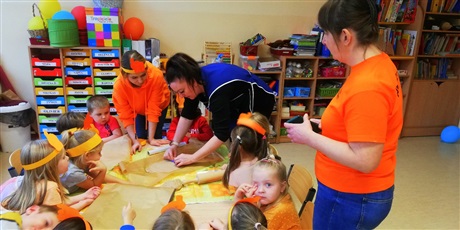 Powiększ grafikę: Na zdjęciu grupa dzieci i 3 osoby dorosłe ubranych  w koszulki koloru pomarańczowego podczas wypieku ciasteczek 