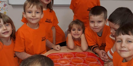Powiększ grafikę: Na zdjęciu dzieci w pomarańczowych koszulkach w sali przedszkolnejj podczas robienia galaretki w kolorze pomarańczowym.