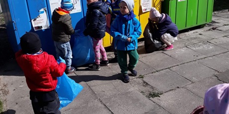 Powiększ grafikę: Na zdjęciu grupa dzieci w wieku przedszkolnym wyrzucająca śmieci do kolorowych pojemników metalowych.