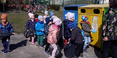 Powiększ grafikę: Na zdjęciu grupa dzieci w wieku przedszkolnym wyrzucająca śmieci do kolorowych pojemników metalowych.