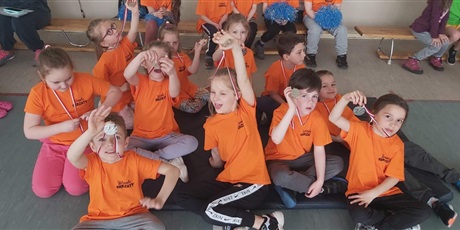 Powiększ grafikę: Na zdjęciu grupa dzieci uśmiechniętych ze srebrnymi medalami w dłoni siedzących na podłodze w sali gimnastycznej.