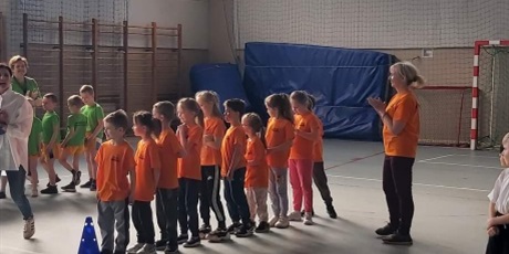 Powiększ grafikę: Na zdjęciu dzieci w wieku przedszkolnym w kolorowych koszulkach stojące w rzędzie na sali gimnastycznej.