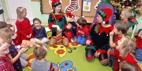 Powiększ grafikę: Na zdjęciu przedstawione są dzieci siedzące na dywanie wśród dwóch pan  Elfów.W tle widać choinkę i dekoracje świąteczne.