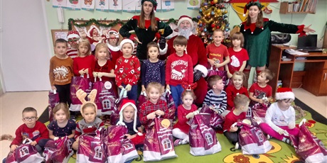 Powiększ grafikę: Na zdjęci grupa dzieci w wieku przedszkolnym w czerwonych strojach i ozdobach mikołajkowych na głowach.Przed sobą na podłodze dzieci trzymają prezenty od Mikołaja,który siedzi wsród dzieci z dwoma Elfami.