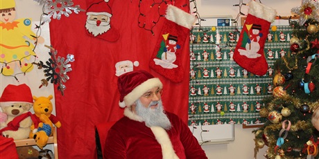 Powiększ grafikę: Na zdjęciu Mikołaj z siwą brodą na tle świątecznej dekoracji i choinki
