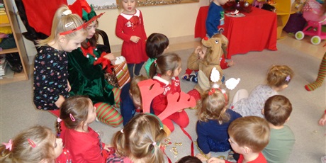 Powiększ grafikę: Na zdjęciu przedstawione są dzieci siedzące na dywanie wśród dwóch pan  Elfów.W tle widać choinkę i dekoracje świąteczne.