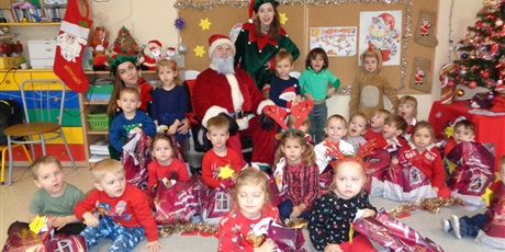 Powiększ grafikę: Na zdjęci grupa dzieci w wieku przedszkolnym z personelem w czerwonych strojach i ozdobach mikołajkowych na głowach.Przed sobą na podłodze dzieci trzymają prezenty od Mikołaja,który siedzi wsród dzieci z dwoma Elfami.