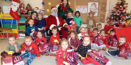 Powiększ grafikę: Na zdjęci grupa dzieci w wieku przedszkolnym z personelem w czerwonych strojach i ozdobach mikołajkowych na głowach.Przed sobą na podłodze dzieci trzymają prezenty od Mikołaja,który siedzi wsród dzieci z dwoma Elfami.