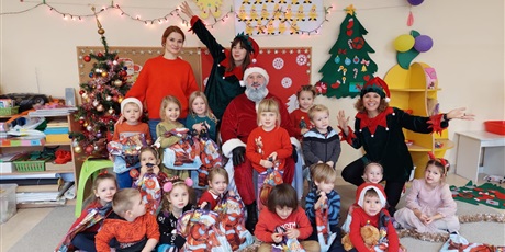 Powiększ grafikę: Na zdjęciu grupa dzieci w wieku przedszkolnym wraz z nauczycielami i Mikołajem w strojach koloru czerwonego i czerwonych czapkach.