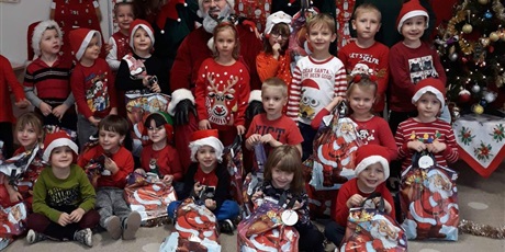 Powiększ grafikę: Na zdjęciu grupa dzieci w wieku przedszkolnym wraz z nauczycielami i Mikołajem w strojach koloru czerwonego i czerwonych czapkach.