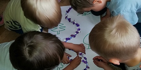 Powiększ grafikę: Na zdjęciu dzieci w wieku przedszkolnym układające koorowe kamyki  na białym papierze po namalowanej lini.