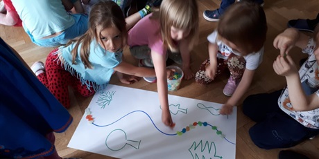 Powiększ grafikę: Na zdjęciu dzieci w wieku przedszkolnym układające koorowe kamyki  na białym papierze po namalowanej lini.