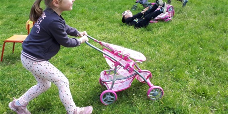Powiększ grafikę: Na zdjęciu dziecko w ieku przedszkolnym biegnące z wózkiem dla lalek po zielonej łące.