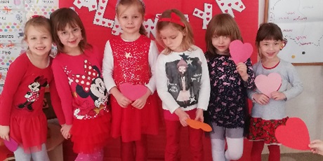 Powiększ grafikę: Na zdjęciu dziewczynki w ieku przedszkolnym w ubraniach koloru czerwonego na tle dekoracji walentynkowej.