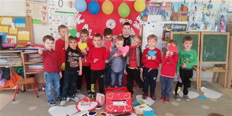 Powiększ grafikę: Na zdjęciu dzieci w wieku przedszkolnym w ubraniach koloru czerwonego na tle dekoracji walentynkowej. Na podłodze stoi pudło poczta walentynkowa oraz listy i laurki walentynkowe.