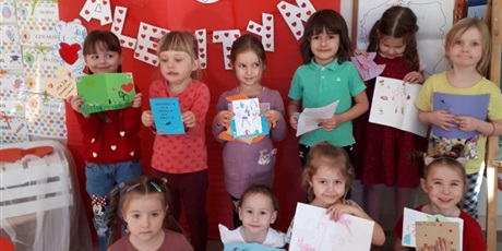 Powiększ grafikę: Na zdjęciu dzieci w wieku przedszkolnym w ubraniach koloru czerwonego na tle dekoracji walentynkowej. Na podłodze stoi pudło poczta walentynkowa oraz listy i laurki walentynkowe.
