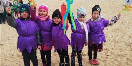 Powiększ grafikę: Na zdjęciu grupa dzieci w wieku przedszkolnym w fioletowych strojach z kwiatkami na plaży witająca wiosnę