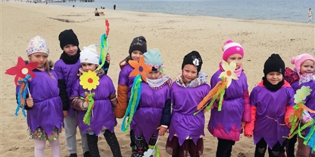 Powiększ grafikę: Na zdjęciu grupa dzieci w wieku przedszkolnym w fioletowych strojach z kwiatkami na plaży witająca wiosnę