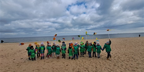 Powiększ grafikę: Na zdjęciu grupa dzieci w wieku przedszkolnym w strojach zielonych z kwiatkami na plaży witająca wiosnę.