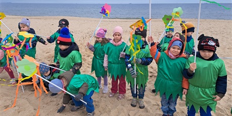 Powiększ grafikę: Na zdjęciu grupa dzieci w wieku przedszkolnym w strojach zielonych z kwiatkami na plaży witająca wiosnę