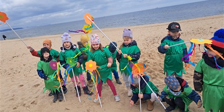 Powiększ grafikę: Na zdjęciu grupa dzieci w wieku przedszkolnym w strojach zielonych z kwiatkami na plaży witająca wiosnę