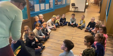 Powiększ grafikę: Pani Przewodnik stoi , za nią znajdują się obrazy przedstawiające dawny Gdańsk. Dzieci siedzą na podłodze. 