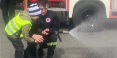 Powiększ grafikę: Na zdjęciu chłopiec ze strażakiem i wężęm gaśniczym tryskającym wodą.