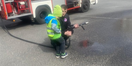 Powiększ grafikę: Na zdjęciu chłopiec ze strażakiem i wężęm gaśniczym tryskającym wodą.