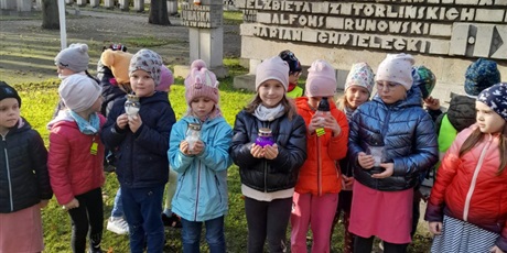 Powiększ grafikę: Na zdjęciu grupa dzieci  w wieku przedszkolnym przed pomnikiem na cmentarzu. ze zniczami w dłoniach