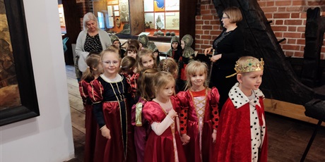 Powiększ grafikę: Na zdjęciu grupa dzieci w wieku przedszkolnym podczas zajęc w muzeum przebrane w czerwone stroje dam dworu, rycerzy  i król