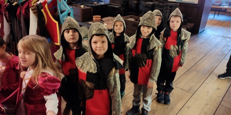 Powiększ grafikę: Na zdjęciu grupa dzieci w wieku przedszkolnym podczas zajęc w muzeum przebrane w czerwone stroje dam dworu, rycerzy