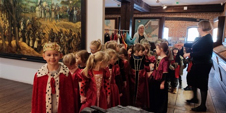 Powiększ grafikę: Na zdjęciu grupa dzieci w wieku przedszkolnym podczas zajęc w muzeum przebrane w czerwone stroje dam dworu, rycerzy