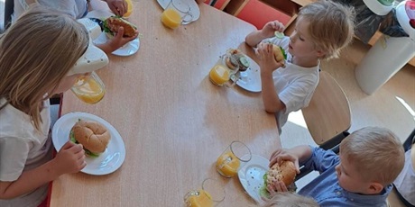 Powiększ grafikę: Na zdjęciu grupa dzieci w wieku przedszkolnym jedząca hamburgery i pijąca sok pomarańczowy.