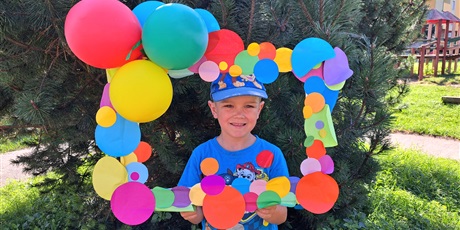 Powiększ grafikę: Na zdjęciu dziecko w wieku przedszkolnym na tle kolorowej ramki ozdobionej papierowymi kropkami i balonami.