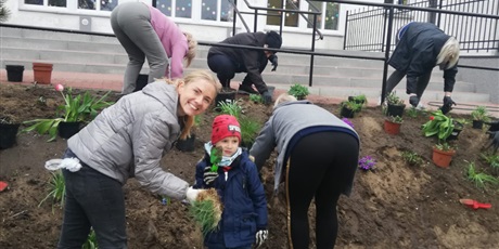 Powiększ grafikę: Na zdjęciu grupa ludzi dorosłych oraz chłopiec w wieku przedszkolnym ze swoją uśmiechniętą mamą  sadzący kwiaty wiosenne w ziemi na rabatce przedszkolnej