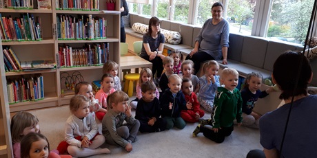 Powiększ grafikę: Przedszkolaki siedzą na dywanie w bibliotece i słuchają opowiadania czytanego przez p. bibliotekarkę.