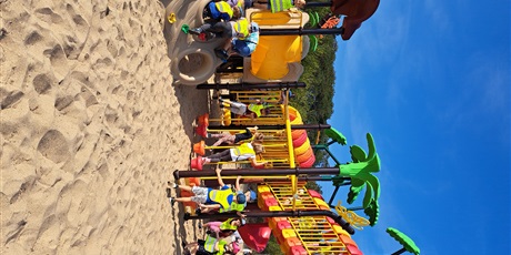 Powiększ grafikę: Na zdjęciu grupa dzieci w wieku przedszkolnym w odblaskowych kamizelkach podczas zabaw na plaży.