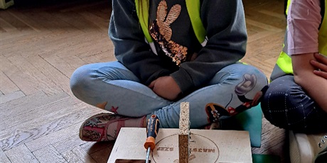 Powiększ grafikę: Na zdjęciu dziecko w wieku przedszkolnym w żóltym kasku kamizelce odblaskowej podczas warszatów stolarskich .W tle młotek stolarski wkrętarka i śrubokręt