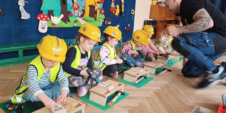 Powiększ grafikę: Na zdjęciu grupa dzieci w wieku przedszkolnym w żółtycha kaskach i kamizelkach odblaskowych podczas warszatów stolarskich