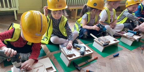 Powiększ grafikę: Na zdjęciu grupa dzieci w wieku przedszkolnym w żółtycha kaskach i kamizelkach odblaskowych podczas warszatów stolarskich