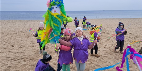 Powiększ grafikę: Na zdjęciu dzieci w kolorowych strojach z papierową panią Wiosną na plaży.